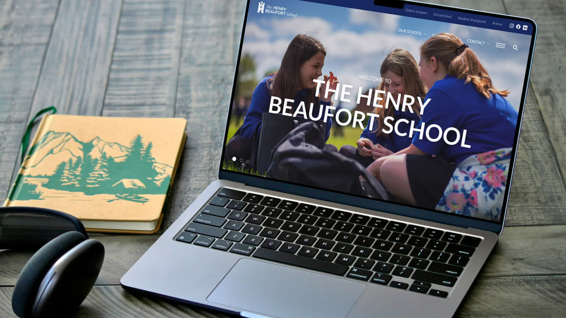 Laptop displaying The Henry Beaufort School homepage on a desk with headphones and a notebook