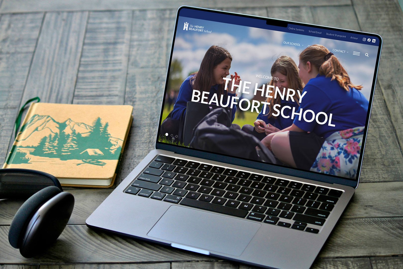 Laptop displaying The Henry Beaufort School homepage on a desk with headphones and a notebook