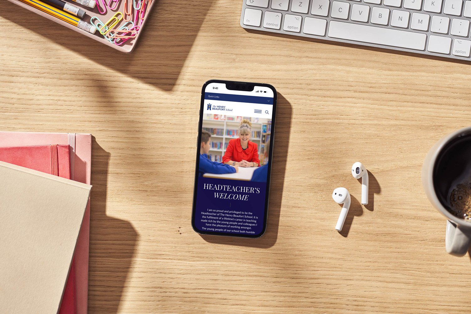 Smartphone showing the school's website on a wooden desk beside stationery, earphones and a coffee cup