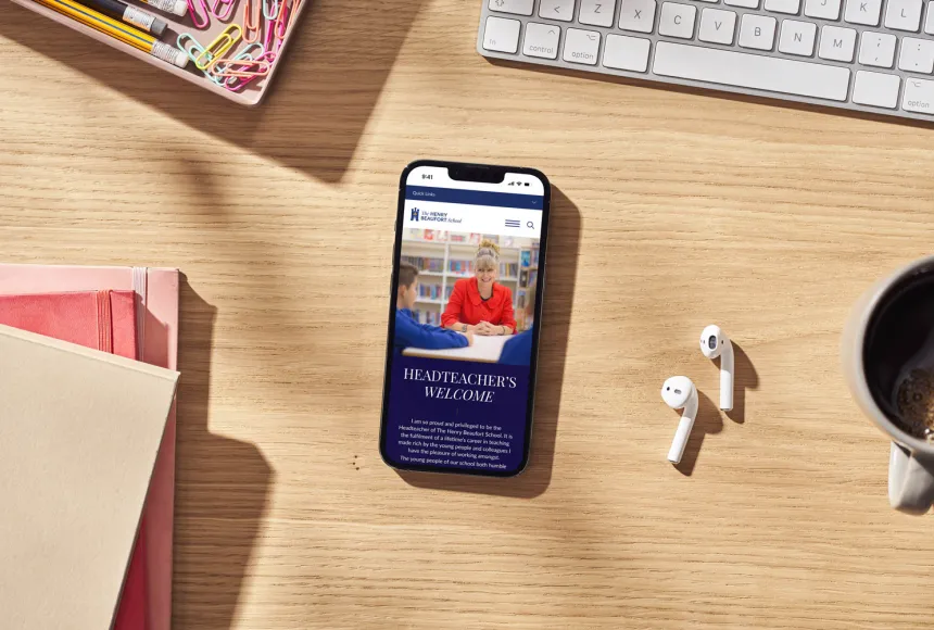 Smartphone showing the school's website on a wooden desk beside stationery, earphones and a coffee cup