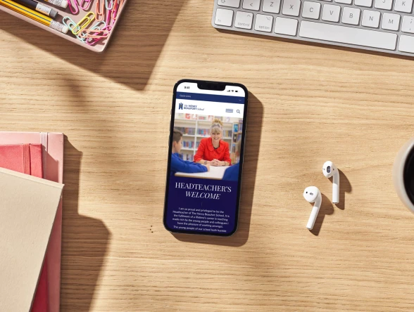 Smartphone showing the school's website on a wooden desk beside stationery, earphones and a coffee cup
