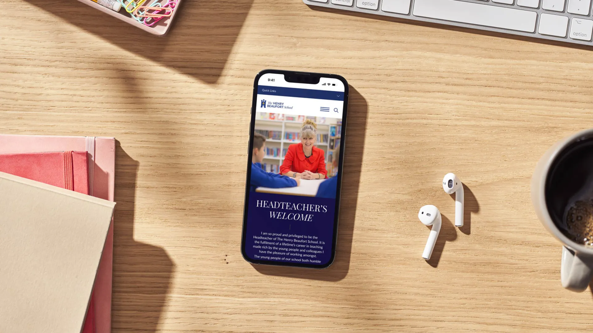 Smartphone showing the school's website on a wooden desk beside stationery, earphones and a coffee cup
