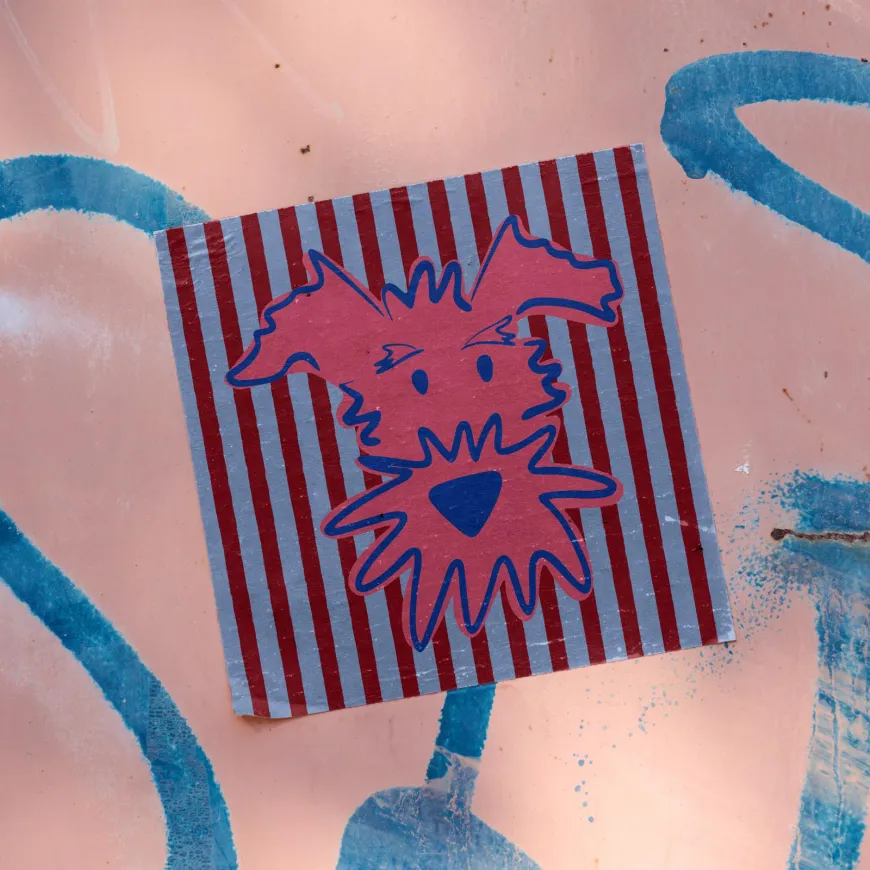 Illustrated dog head in pink and blue on a striped sticker, on a pink graffiti wall.