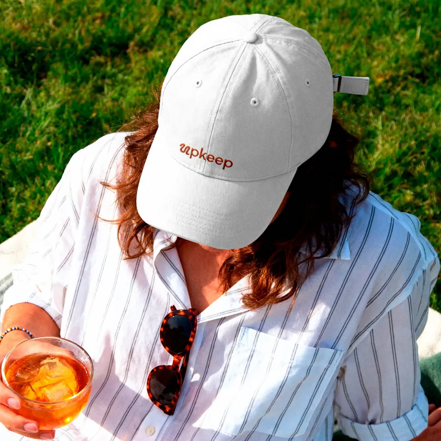 Lady wearing a white striped shirt and a white baseball cap hat with the Upkeep logotype embroidered in orange. Whilst holding a negroni cocktail.