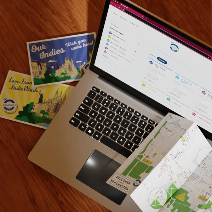Laptop mockup, postcards and Winchester map laid out on a wooden surface.